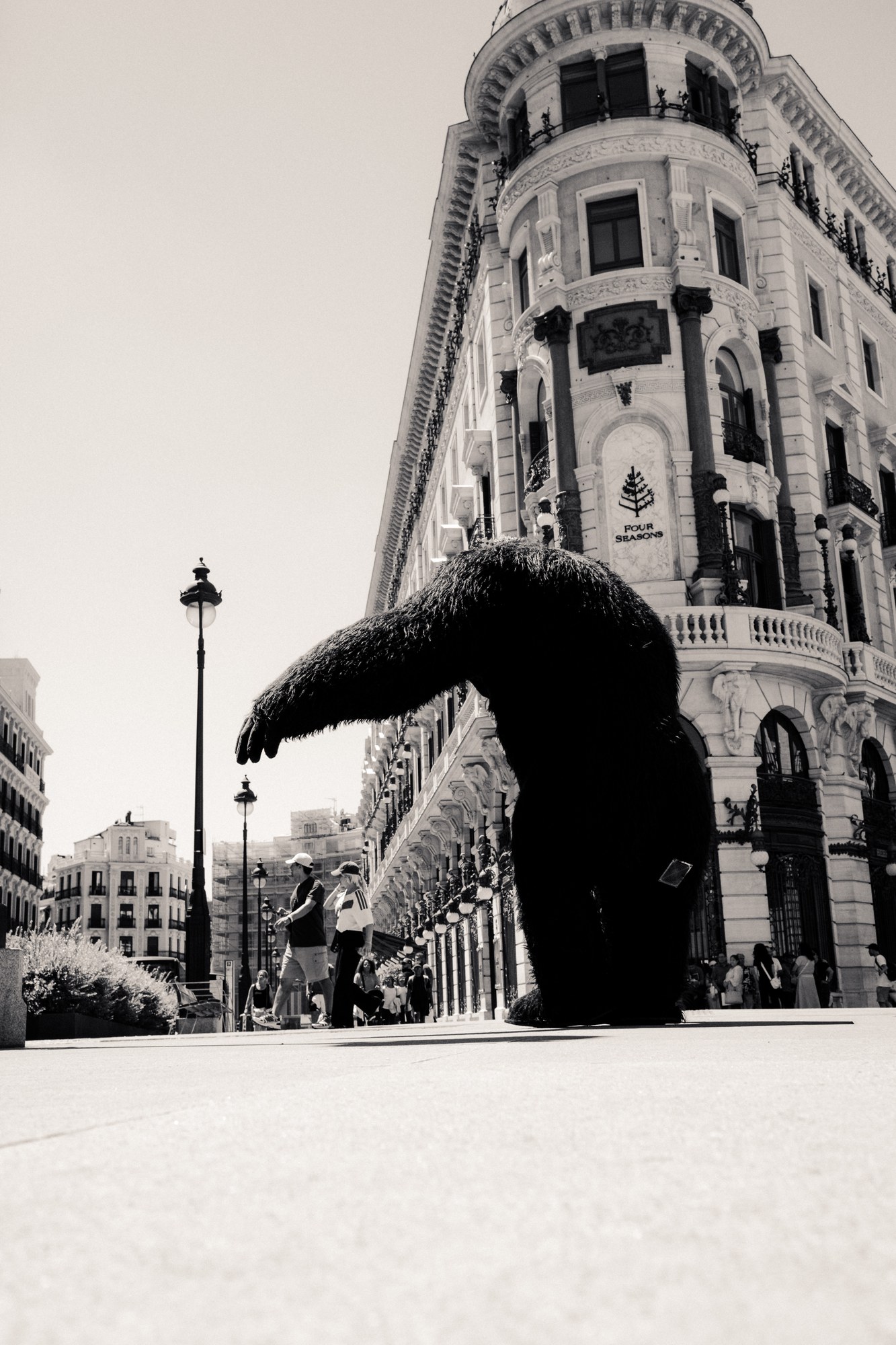 Photographed from ground level in black and white, a huge dark shaggy figure with one arm outstretched stands on an empty square in front of an ornate corner building. Nobody looks at it.