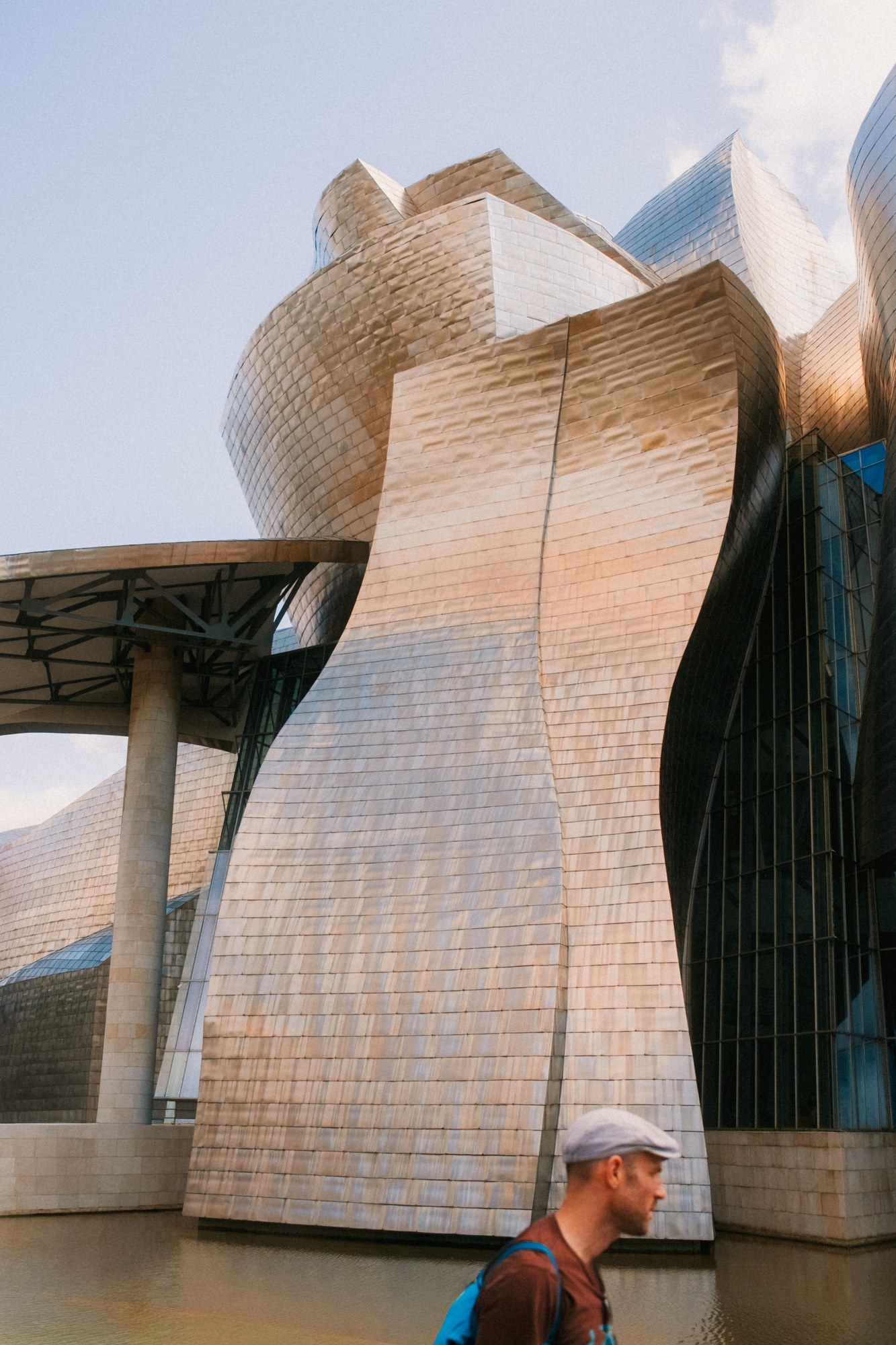 The curved metal panels of the Guggenheim in Bilbao catch the evening light; at the lower edge a man in a flat cap stands in front of them, looking away.