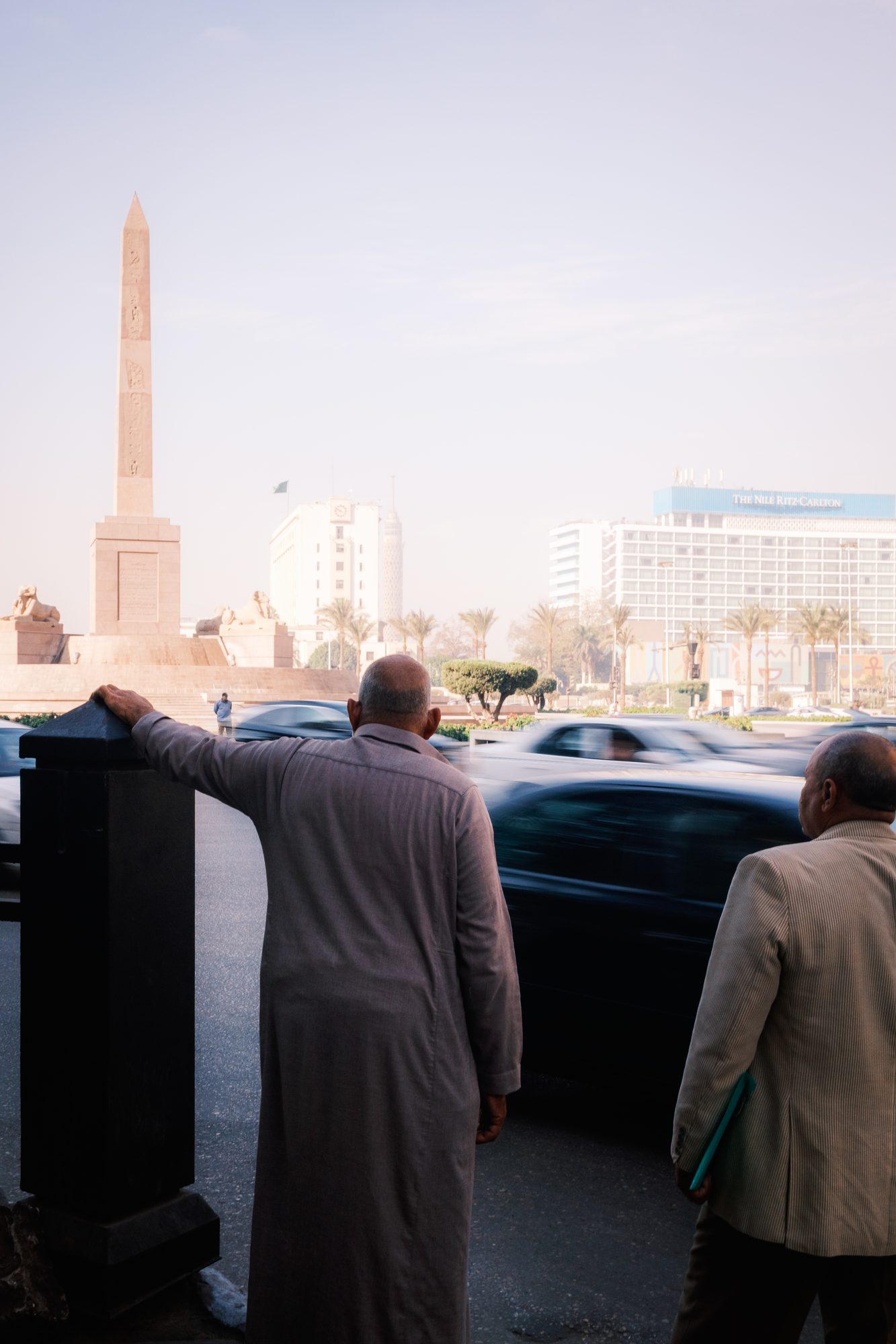 A man in a galabiya stands with his back turned by an obelisk while traffic blurs past in the haze.