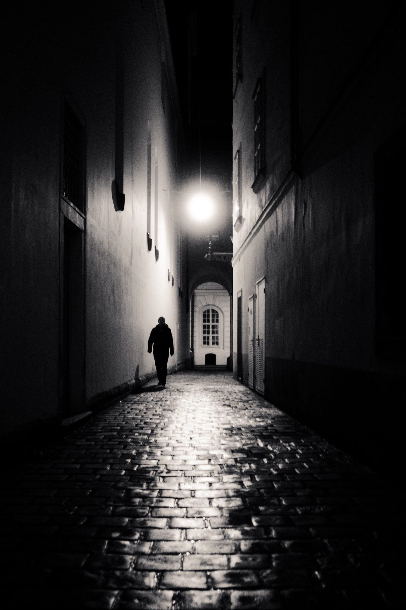 A wet cobblestone lane at night, one street lamp glaring, a figure walking away.