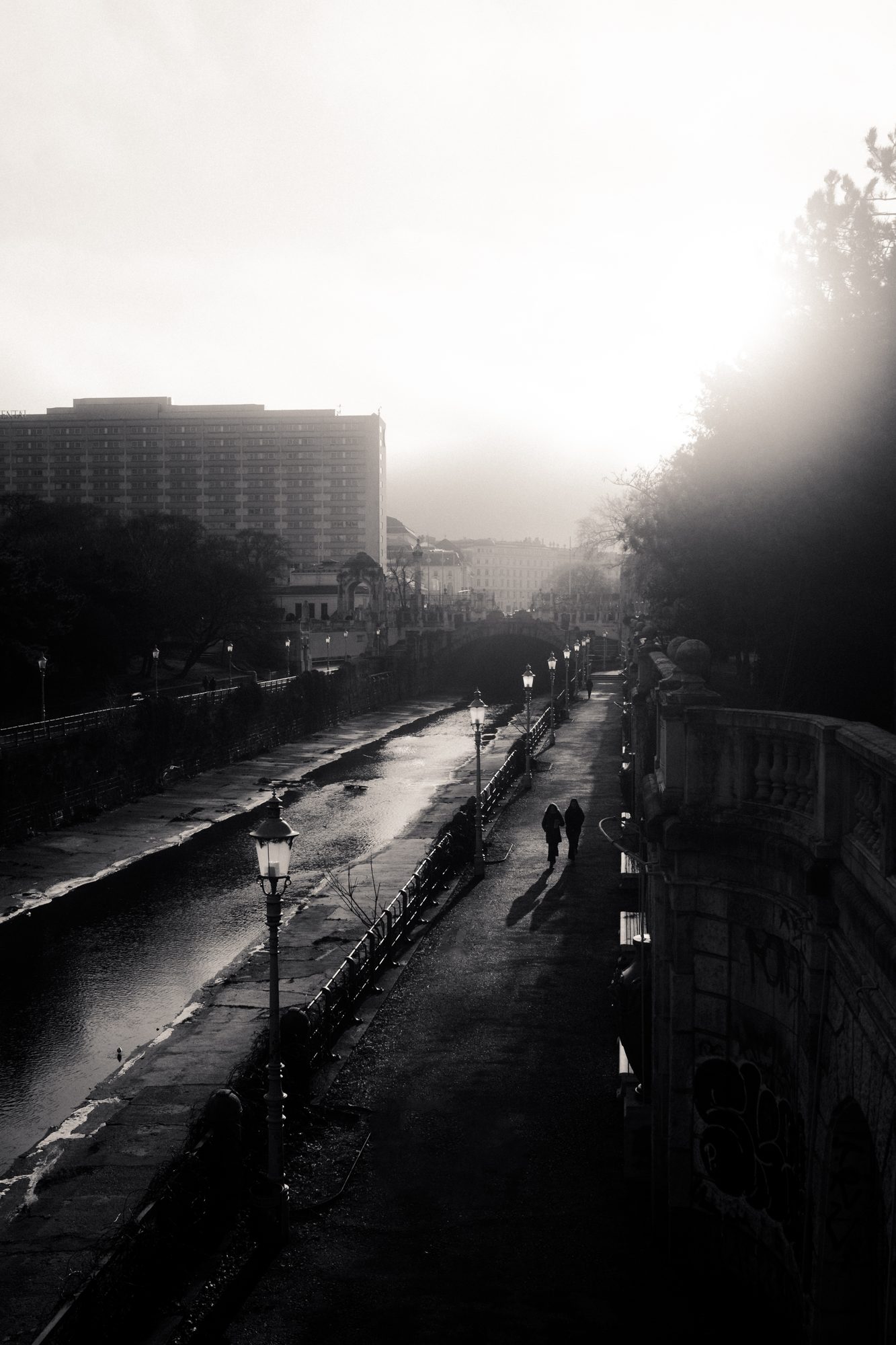 Two figures on a wet river promenade, walking into the morning glare.
