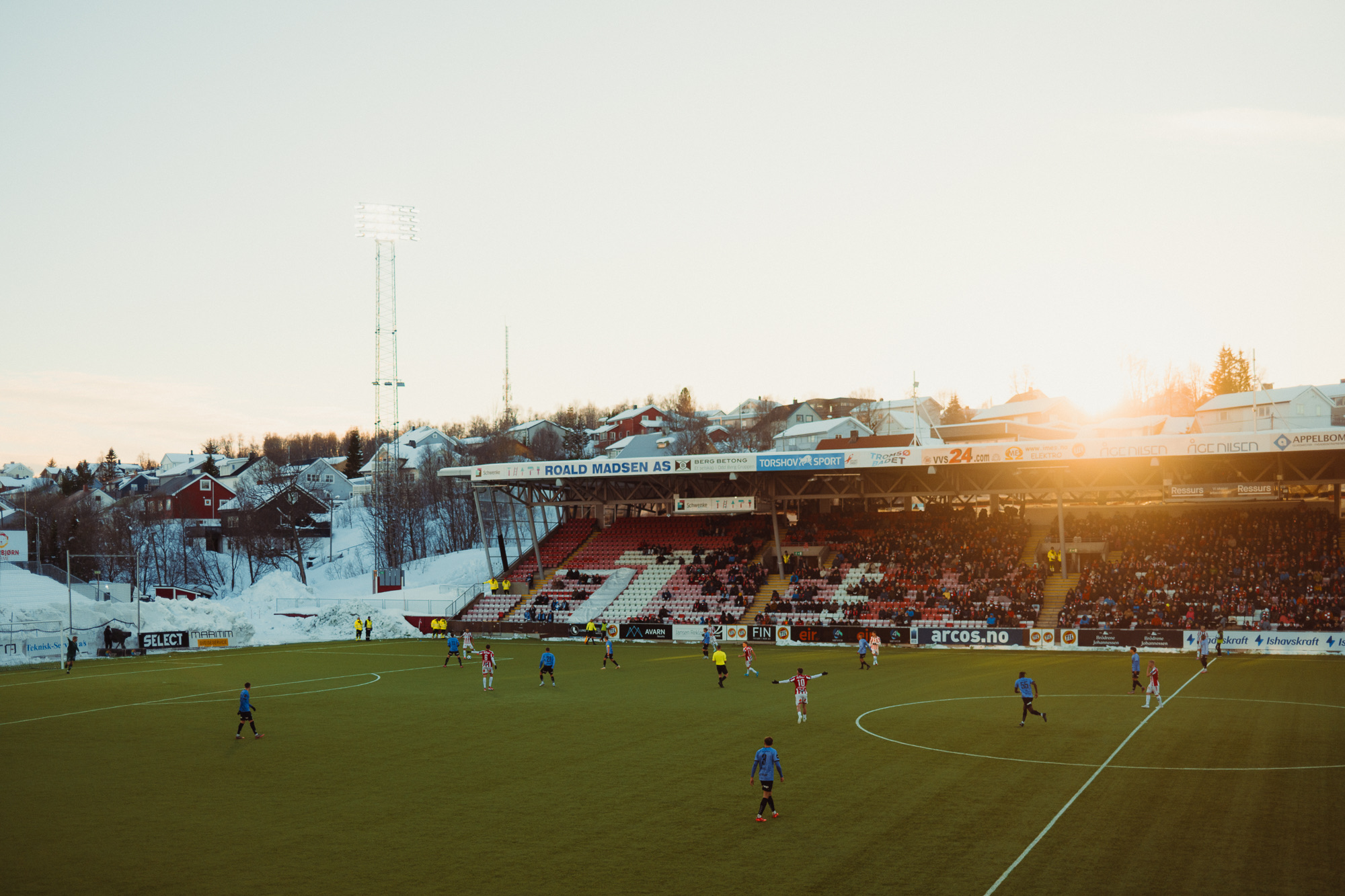 A football match at sundown, small players on a green pitch, snow on the hills behind the stand.
