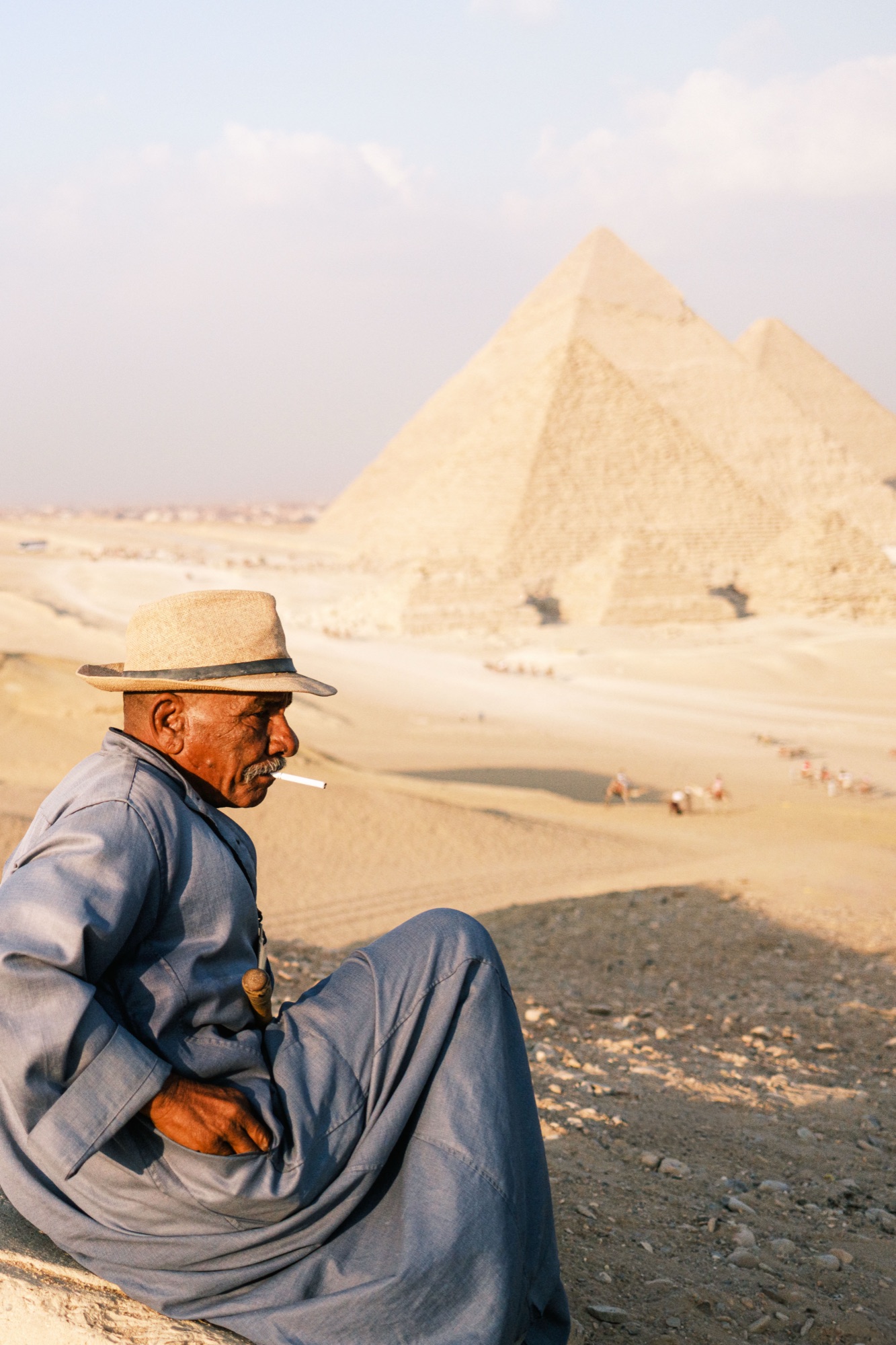 A man in a blue galabiya sits and smokes, the pyramids of Giza soft behind him.