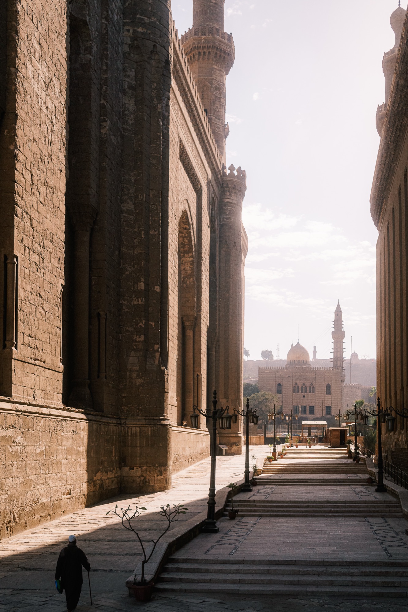 A man with a cane walks in shadow past a monumental mosque wall in old Cairo, morning light.