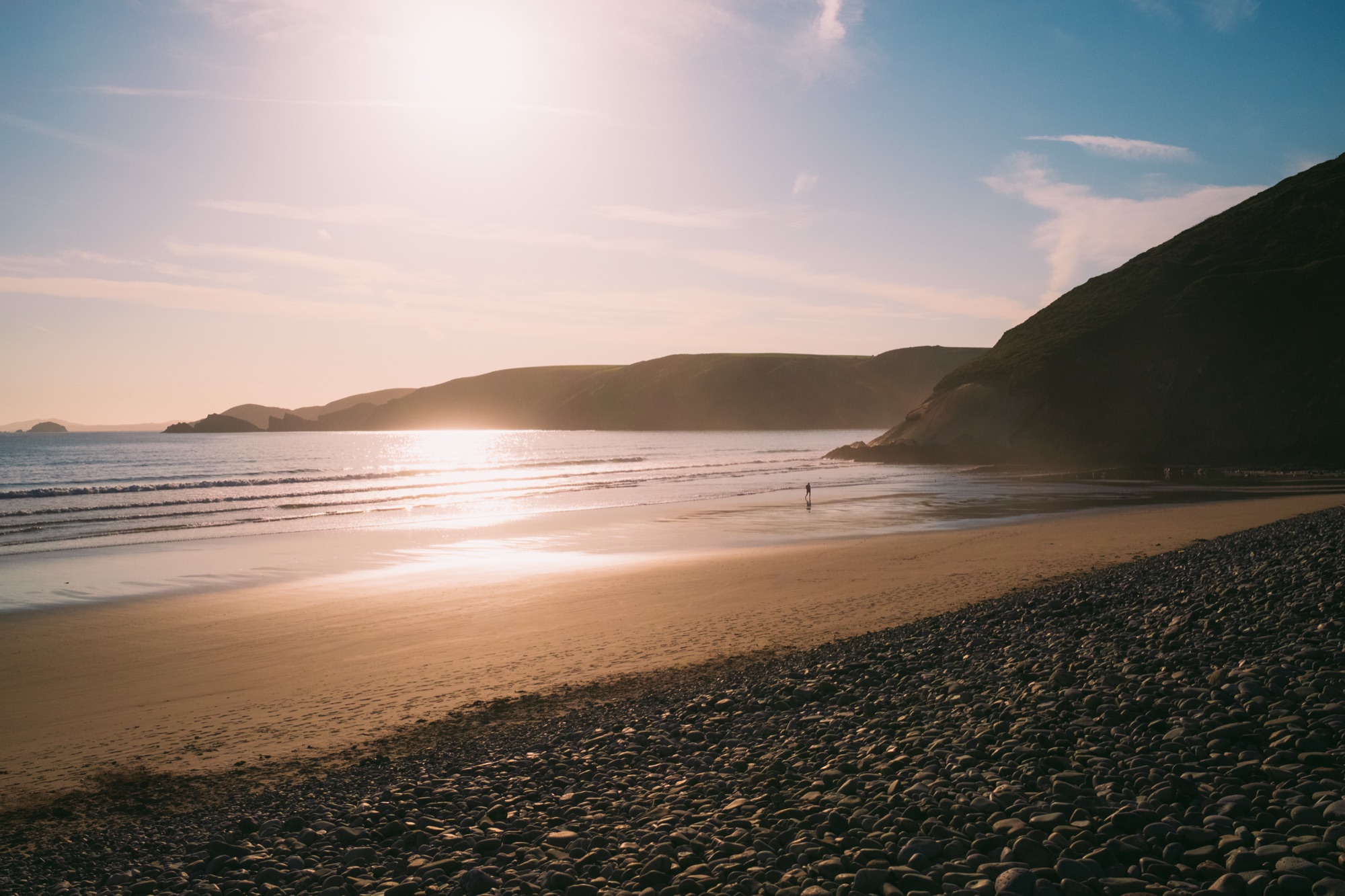 A single figure walks a wide beach at sunset, pebbles in the foreground.