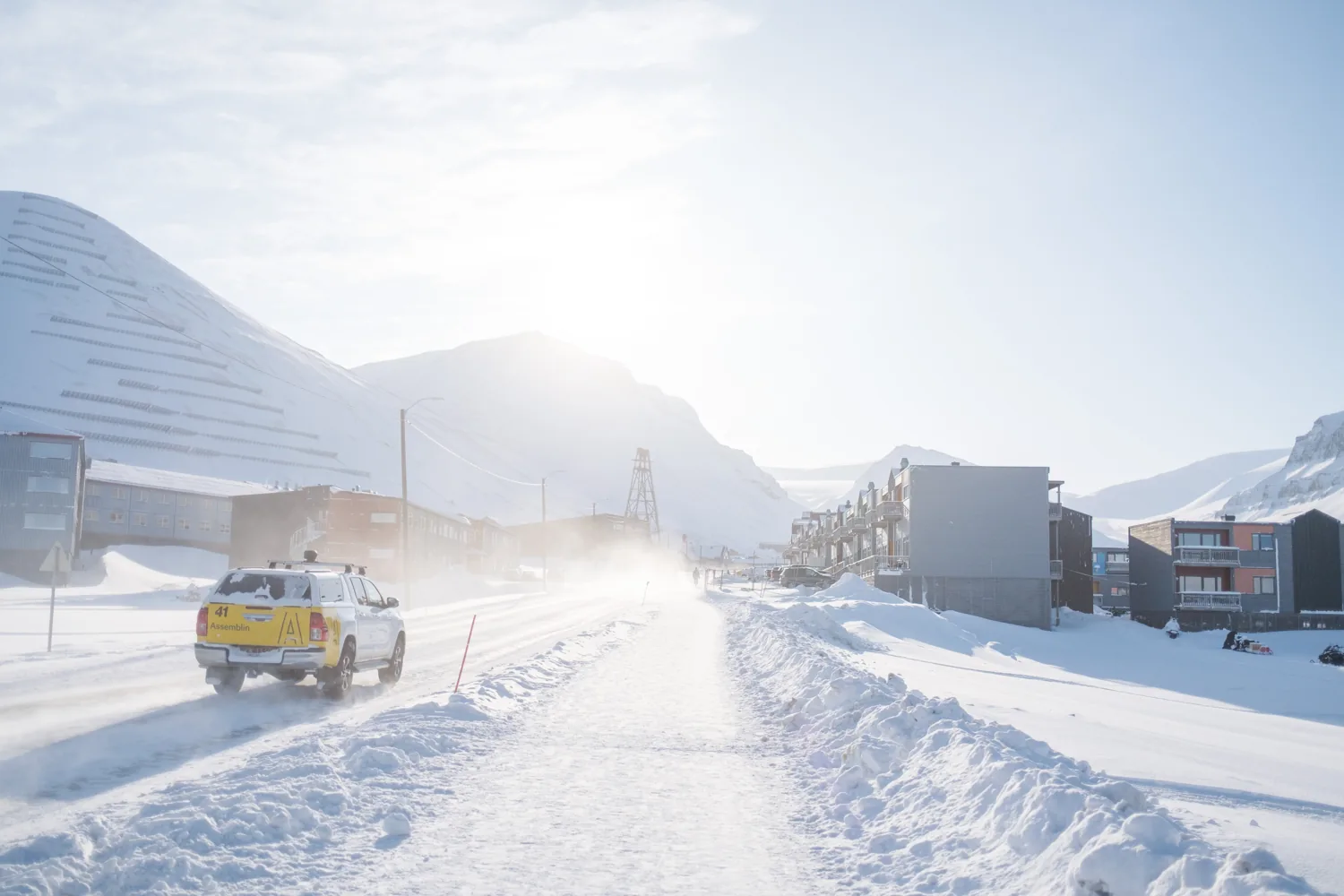 A yellow vehicle drives down a snowy road trailing spray, backlit, town and mountains behind.