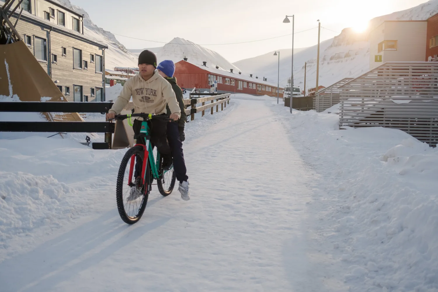 A person cycling along a cleared path through the snowy town, low sun behind the buildings.