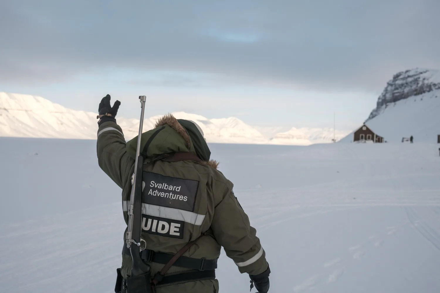 A guide seen from behind raises an arm toward the wide snowfield and mountains.