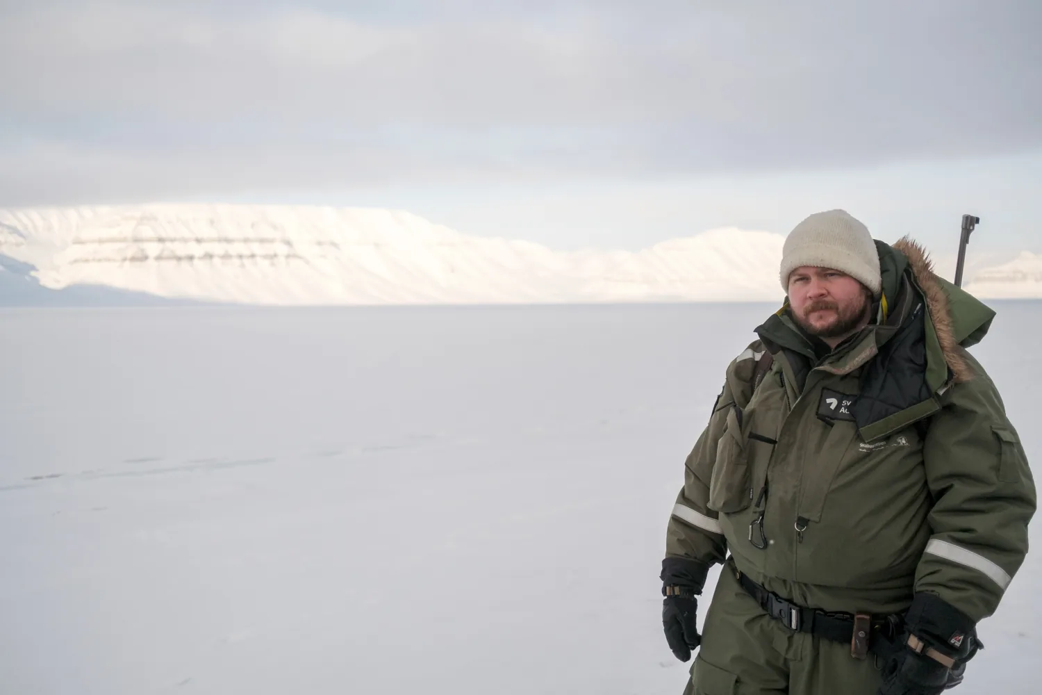 A guide in a heavy parka stands in the snow with a rifle over his shoulder.
