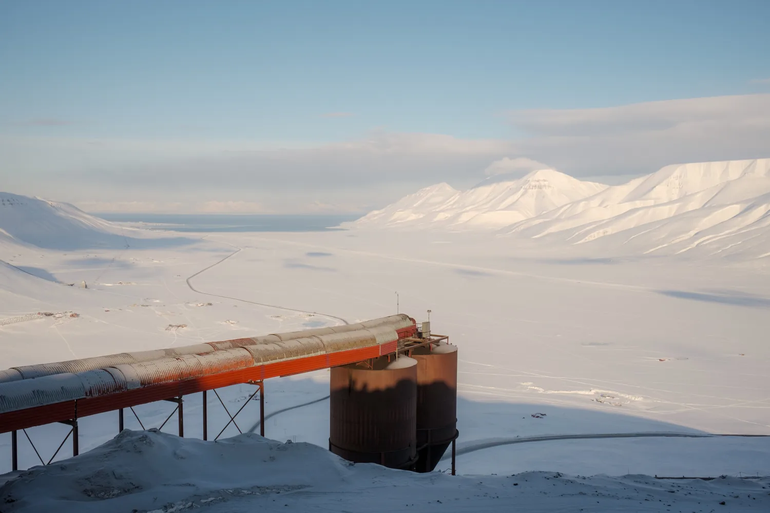 An orange pipeline on stilts crosses the snow, mountains behind.
