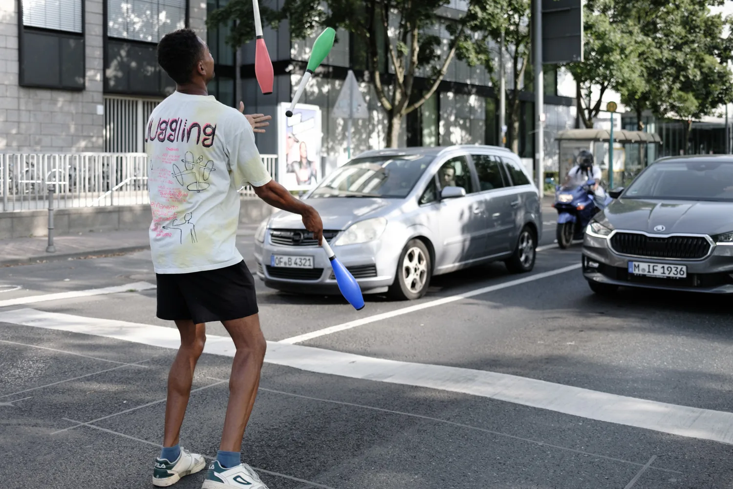 A young man in a 'Juggling' shirt juggles at the edge of the road.