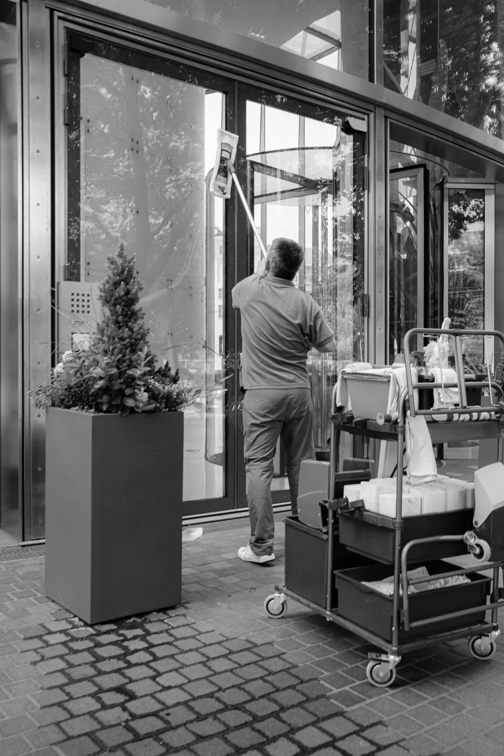 A cleaner with a cart works at a glass entrance.