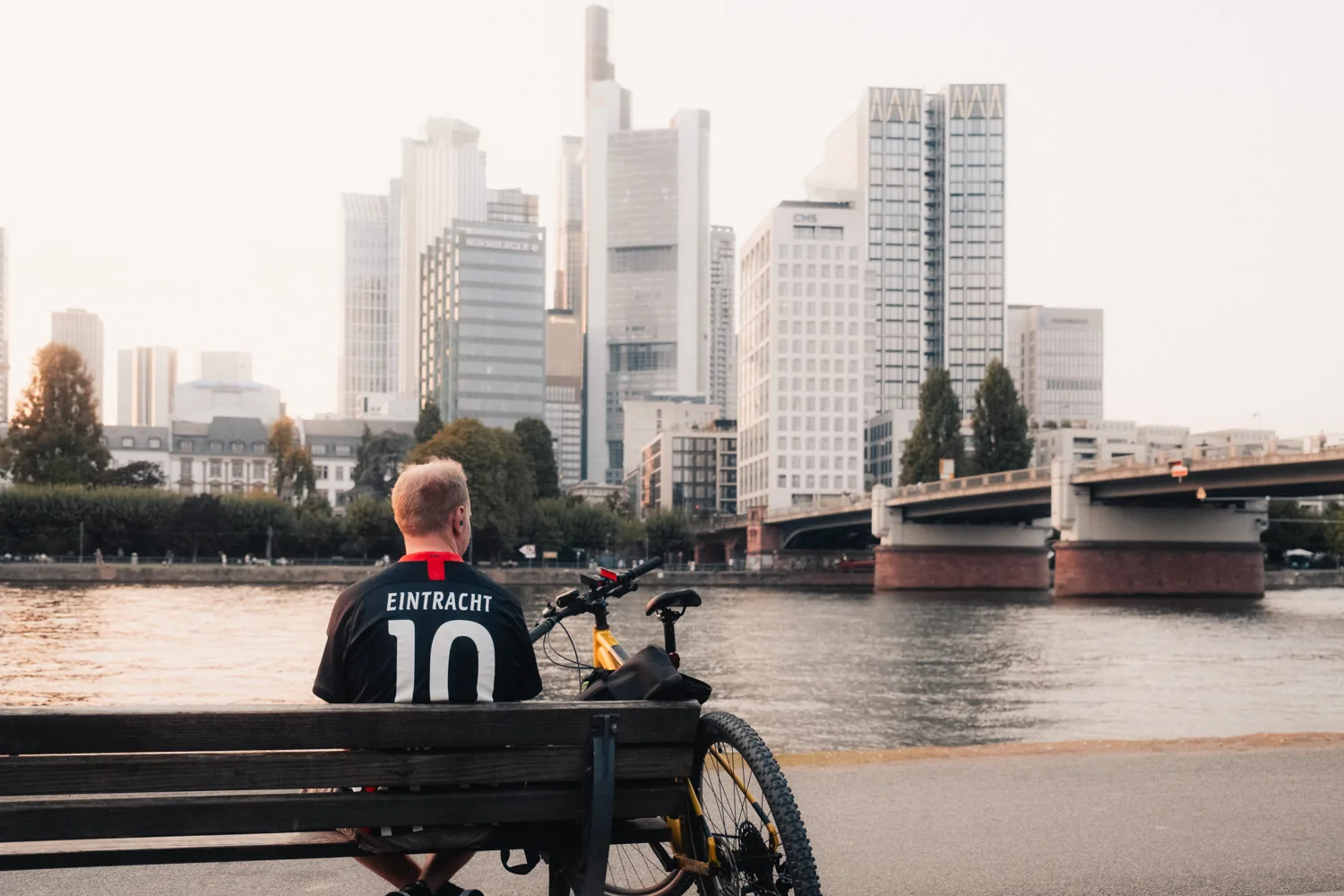 A person in an Eintracht number-ten shirt sits on a bench facing the skyline across the river.