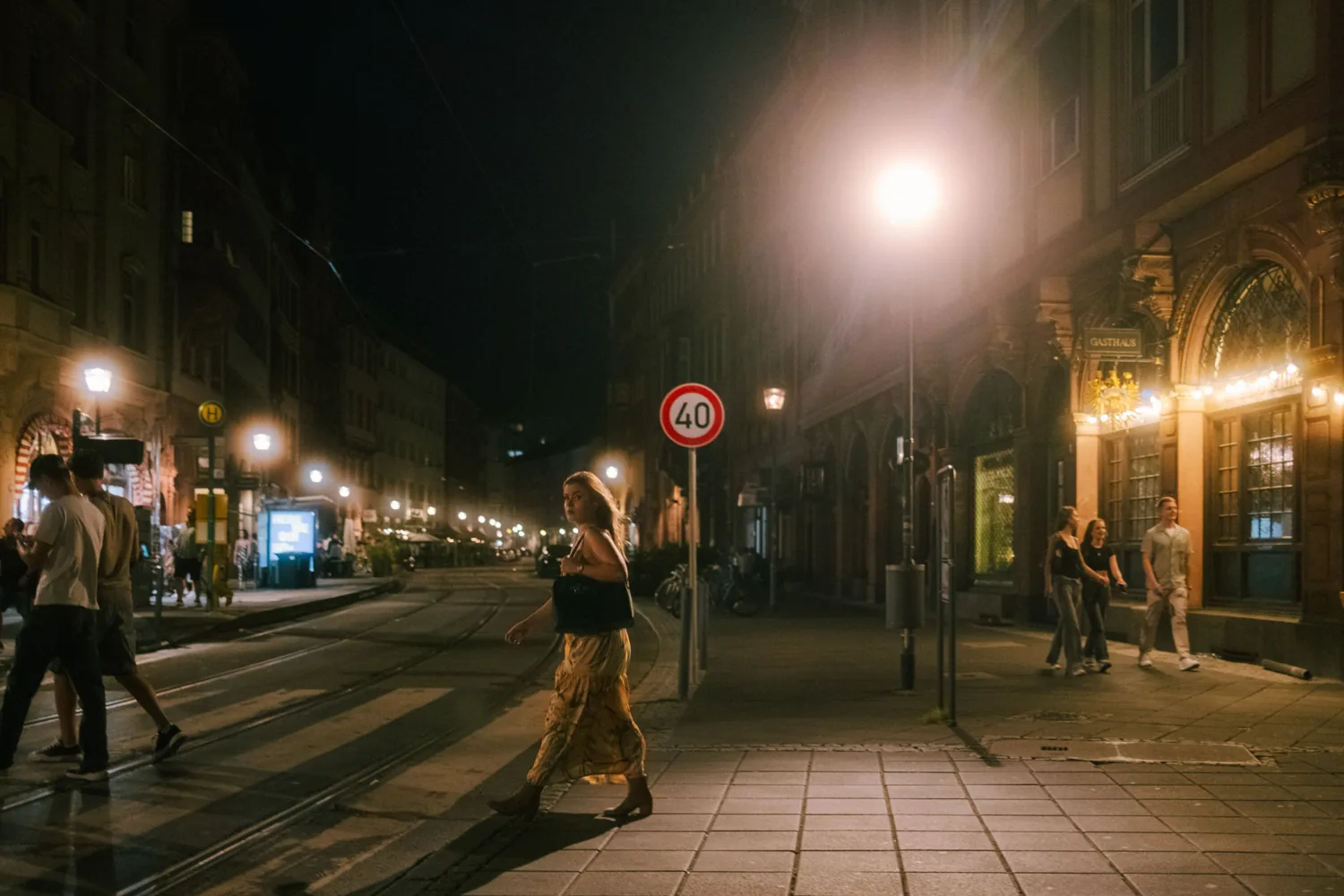 A woman in a yellow dress crosses the tram tracks at night under a street lamp.