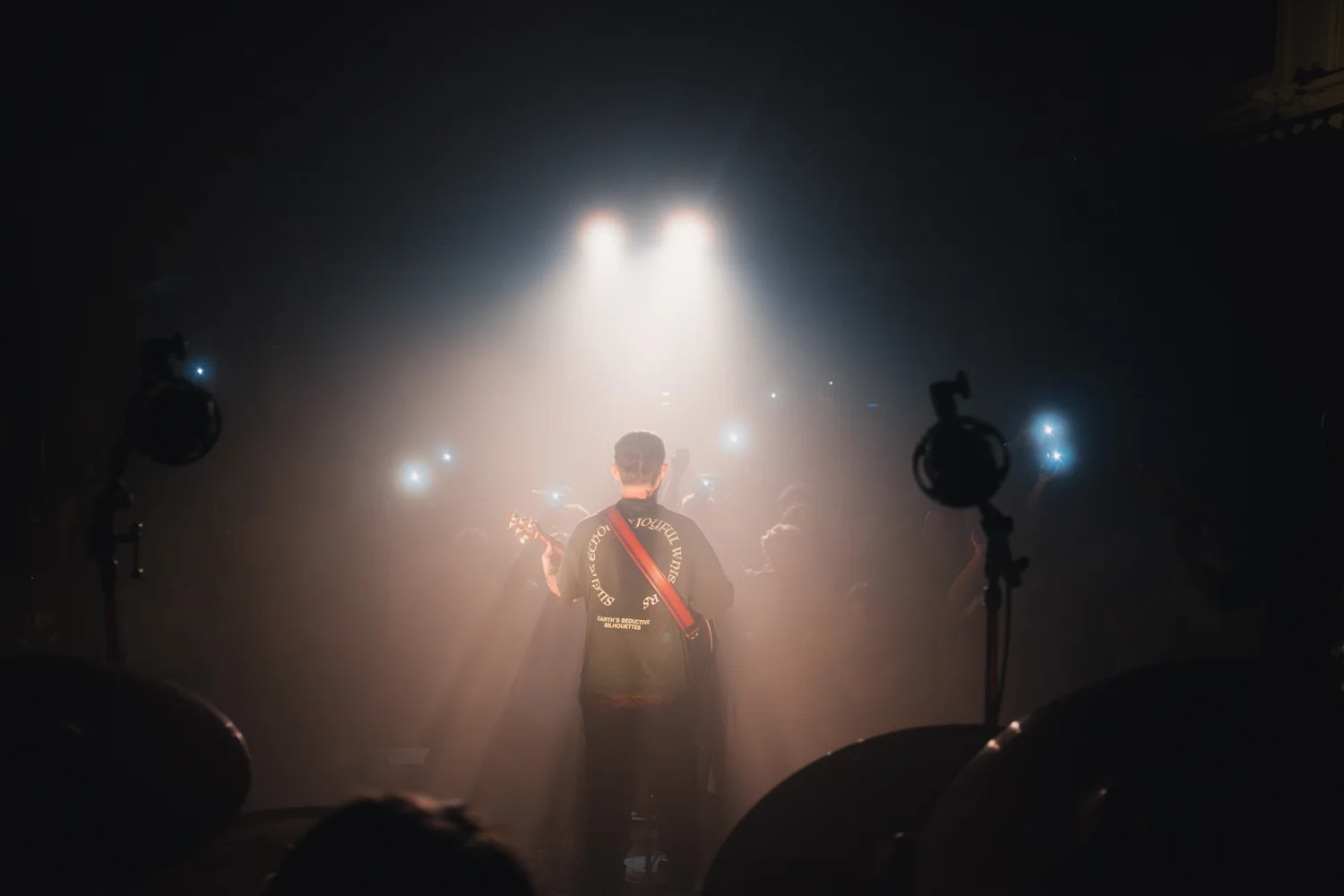 A bass player in silhouette on stage against fog and the phone lights of the crowd.