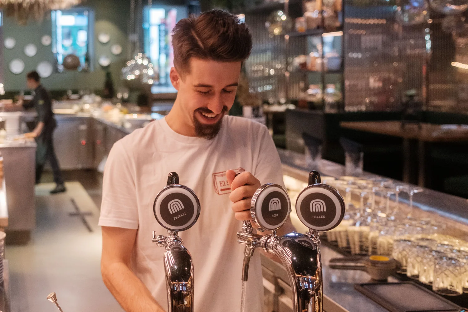 A smiling bartender pours a drink at the taps of a warmly lit bar.